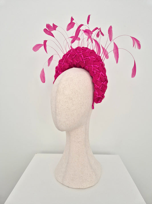 A raspberry pink fascinator headpiece with a textured woven crown and long feather plumes, displayed on a cream-colored felt mannequin head against a plain white background. side view