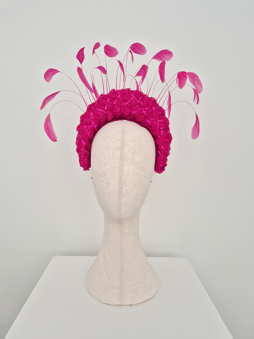 A raspberry pink straw braid fascinator headpiece with long, slender pink feathers like accents, displayed on a cream-colored mannequin head against a light gray background.