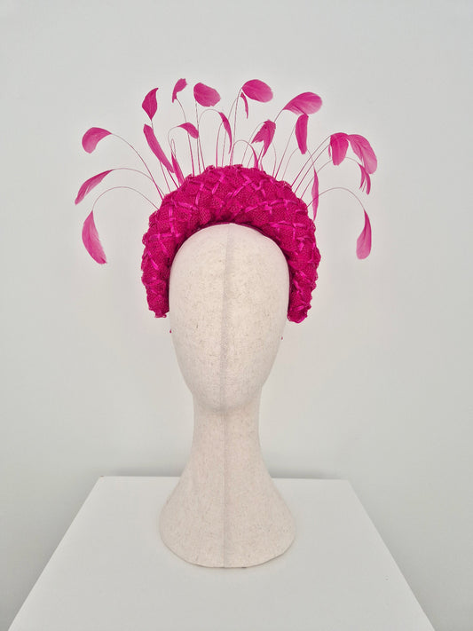 A raspberry pink straw braid fascinator headpiece with long, slender pink feathers like accents, displayed on a cream-colored mannequin head against a light gray background.