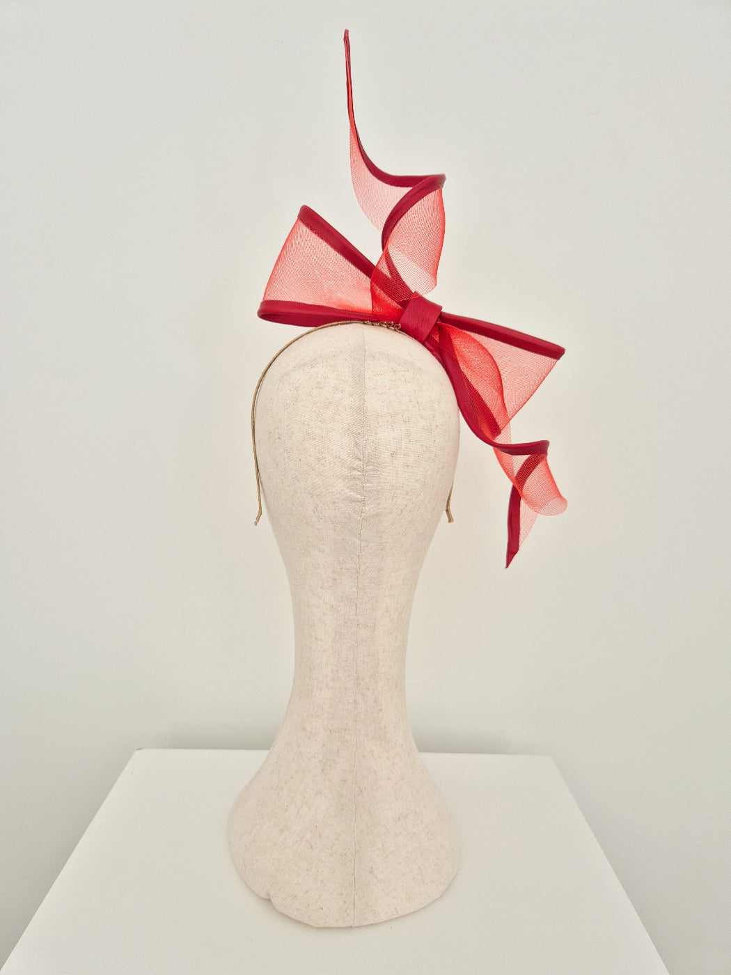 A red crinoline fascinator headpiece with a large bow and flowing ribbon tails, displayed on a cream-colored felt mannequin head against a plain white studio background.back view