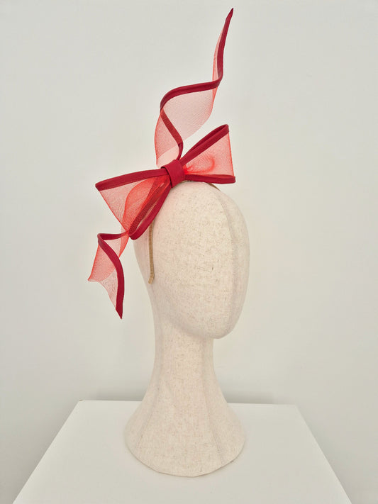 A red crinoline fascinator  with a large bow and sweeping ribbon tails, displayed on a textured off-white mannequin head against a plain light gray background. side view