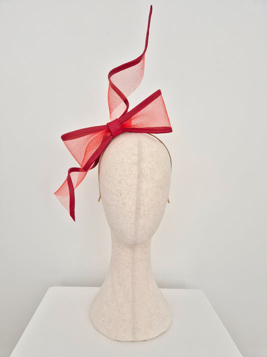 A red crinoline fascinator with a large bow and dramatic upward ribbon tails, displayed on a cream-colored felt mannequin head against a light gray background.