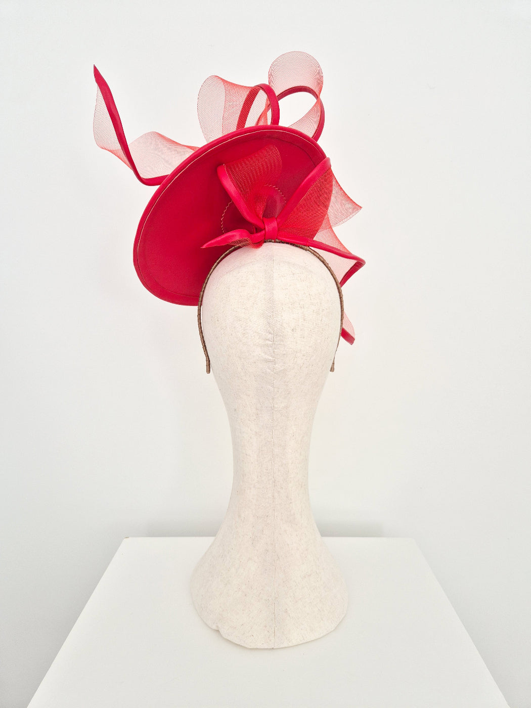 A red leather percher fascinator hat with crinoline loops and a matching bow, displayed on a cream-colored felt mannequin head against a white background.back view