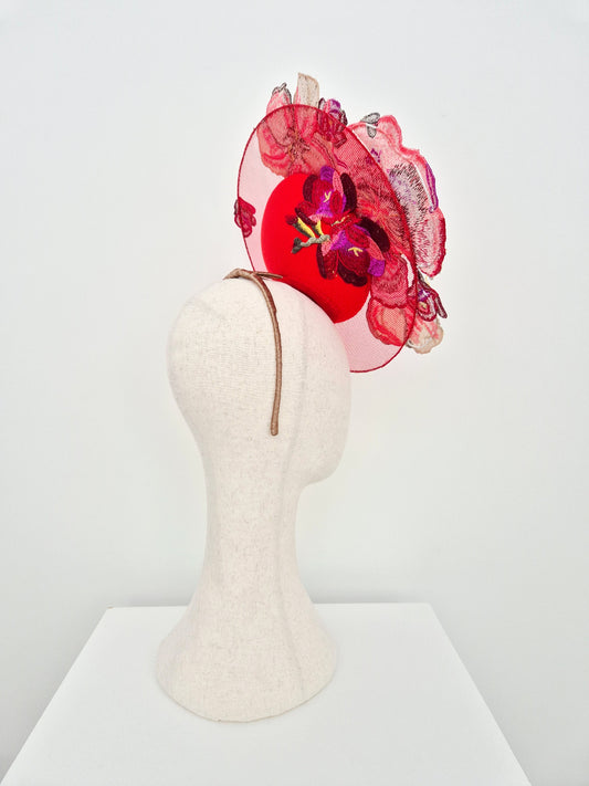 A red fascinator hat with floral embellishments, displayed on a cream-colored felt mannequin head against a white background.side view