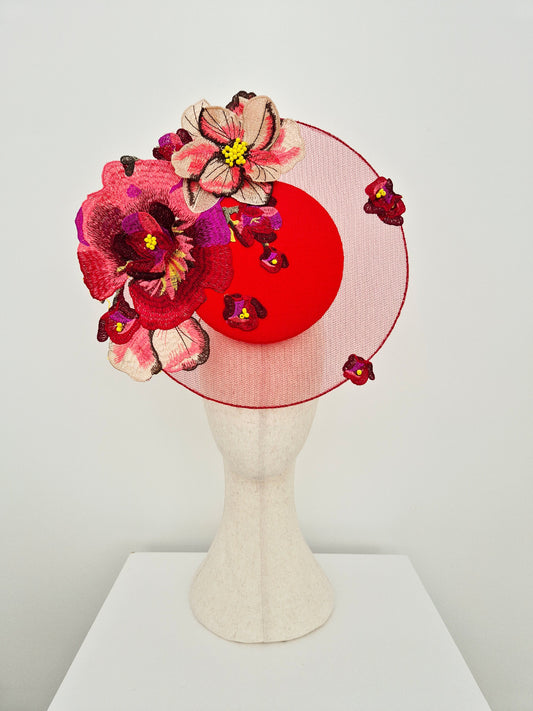 A red satelitte percher headpiece with large embroidered floral decorations in red, pink, and purple, displayed on a cream-colored mannequin head against a light gray background.