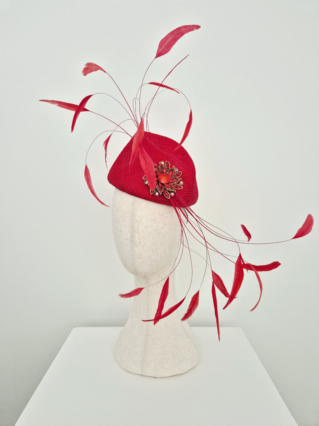 A bright red straw fascinator  displayed on a  mannequin head. The headpiece features a structured red crown with long, flowing red feathers and a decorative red and gold floral embellishment on the front. side view