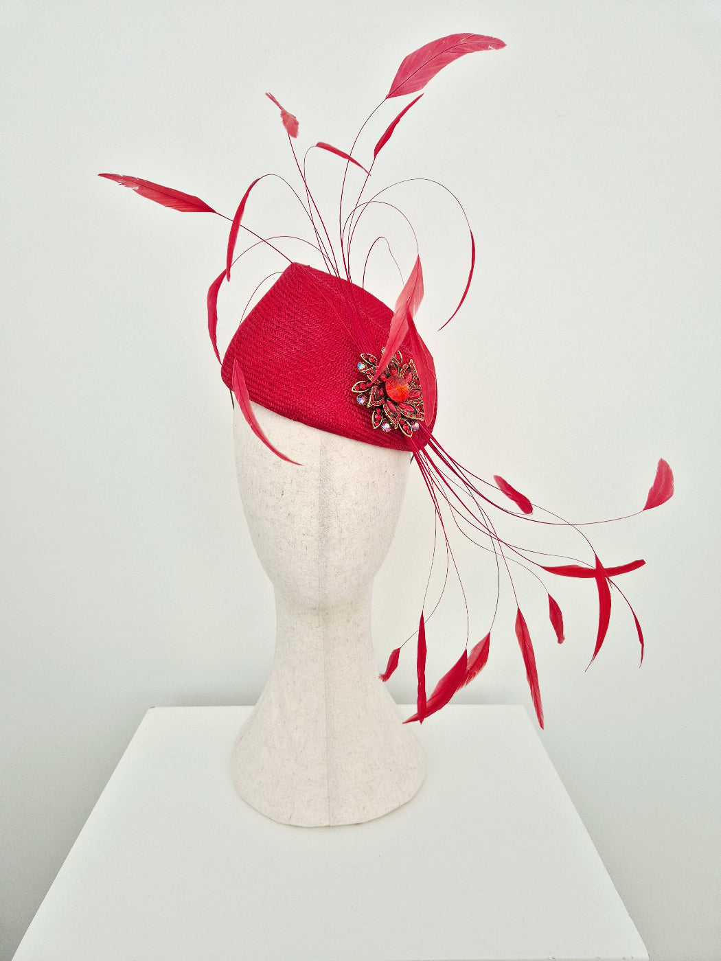 A bright red fascinator  displayed on a cream-colored mannequin head. The hat features a structured red straw base with long, wispy red feathers and a decorative red and metallic floral embellishment on the side.