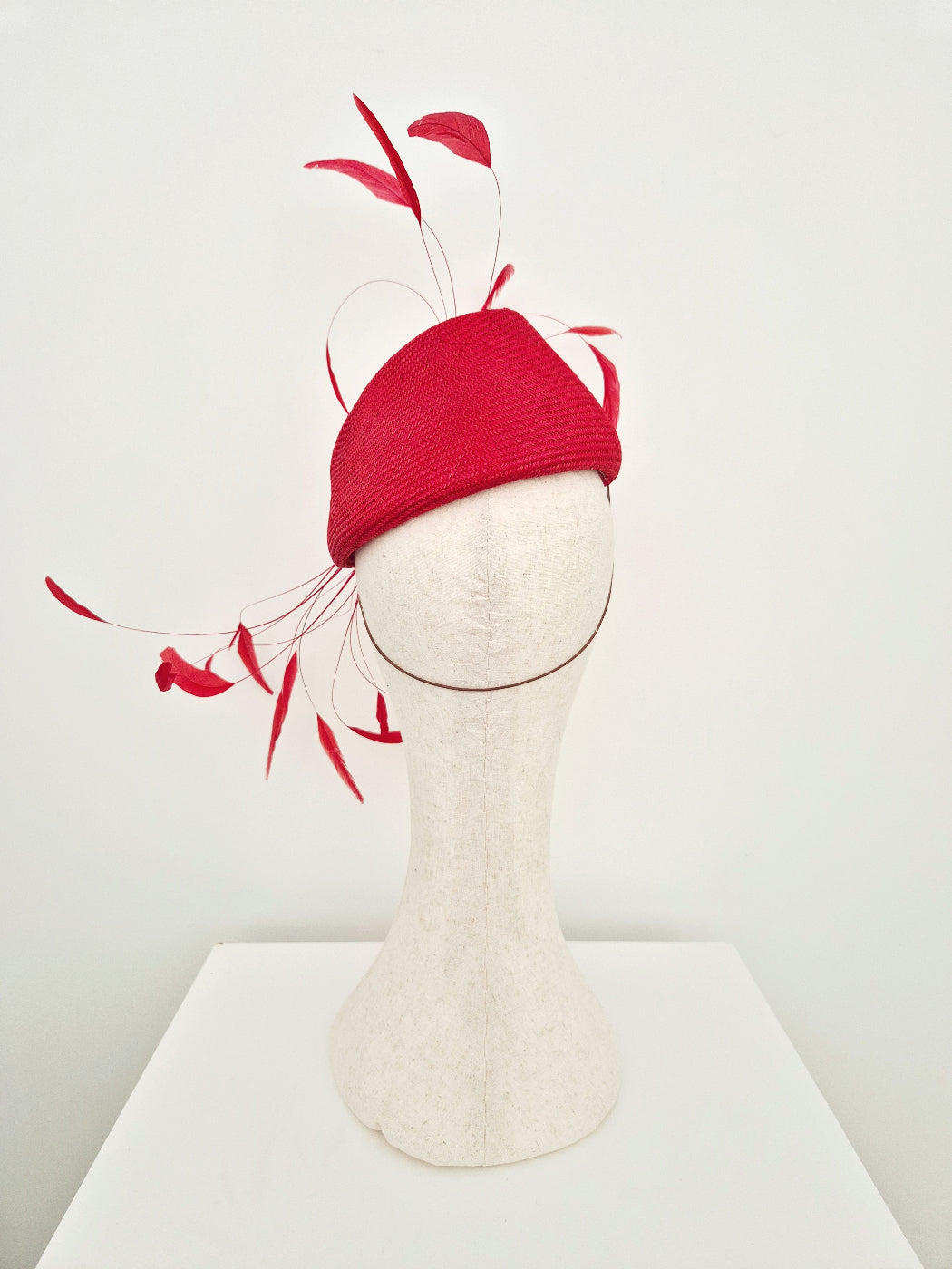 A bright red fascinator hat with long feather-like accents, displayed on a white mannequin head against a clean white background.back view