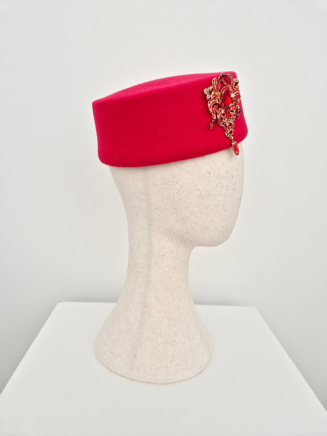 Red pillbox headpiece with decorative brooch on a mannequin head against a white background side view