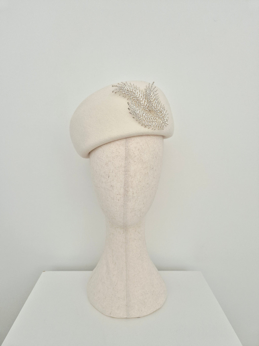 Ivory wool felt beret headpiece with a silver fern rhinestone embellishment on a manniquin head against a white gray background
