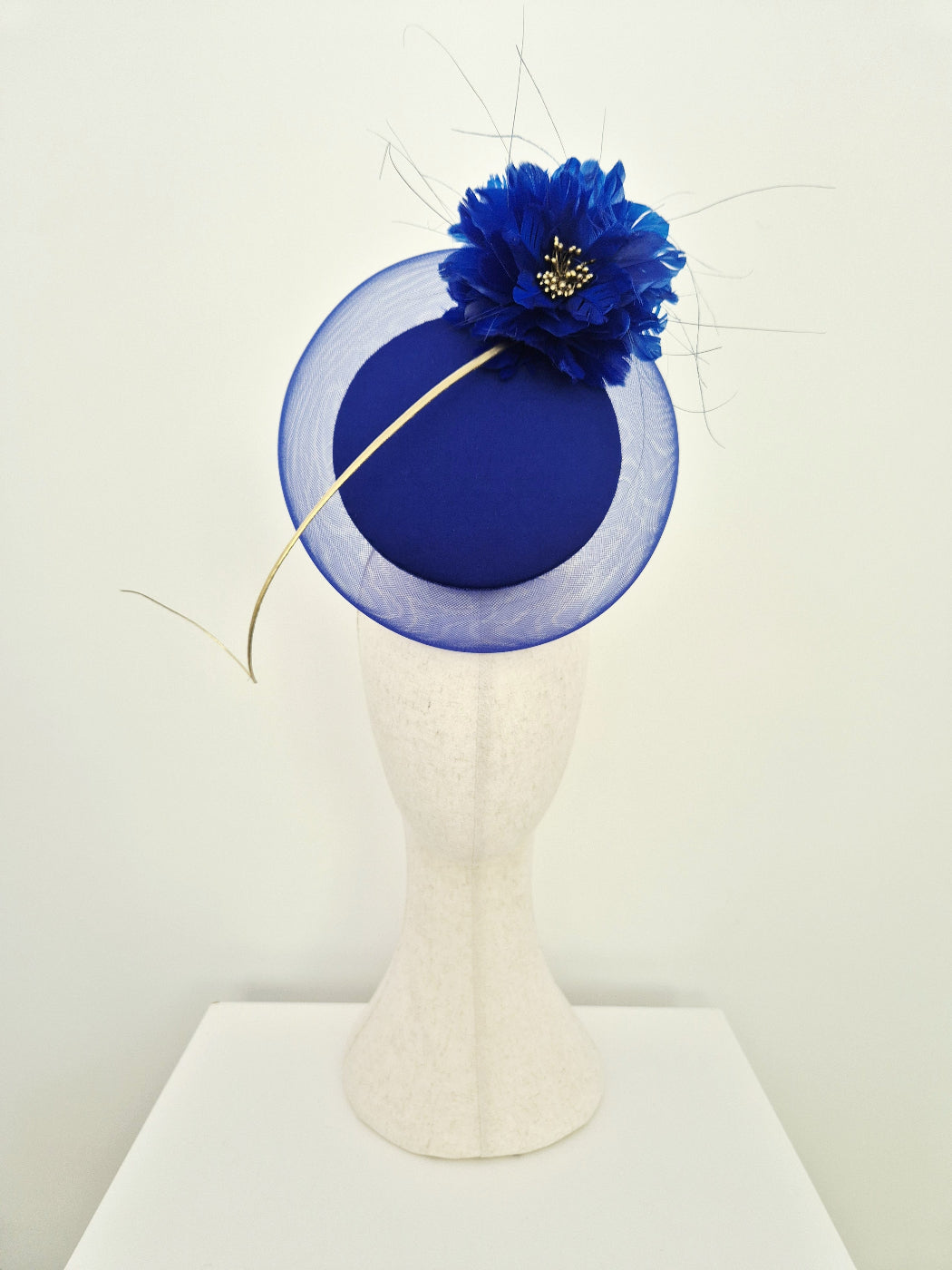 A royal blue percher fascinator  with a large flower accent and gold quill displayed on a textured white mannequin head against a plain off-white background.
