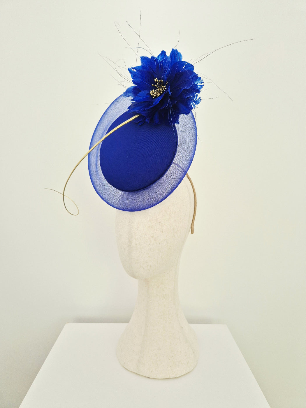 A royal blue fascinator hat with a sheer oval brim, a large blue flower accent, and thin gold quill, displayed on a cream-colored mannequin head against a white background side view