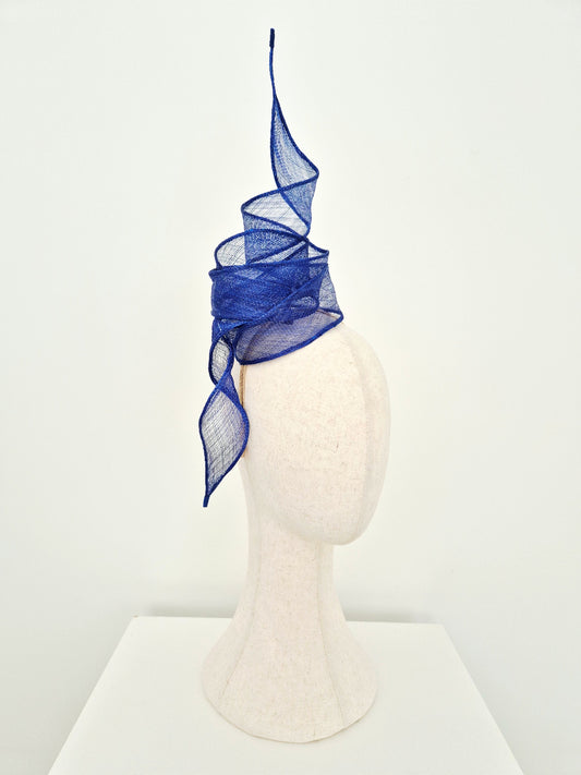 A sculptural royal blue sinamay straw fascinator hat displayed on a cream-colored mannequin head against a clean white background.side view