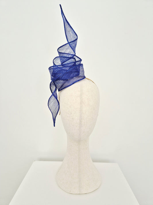 A striking royal blue fascinator made of sinamay straw twisted knot and tails, displayed on a cream-colored felt mannequin head against a clean white background.