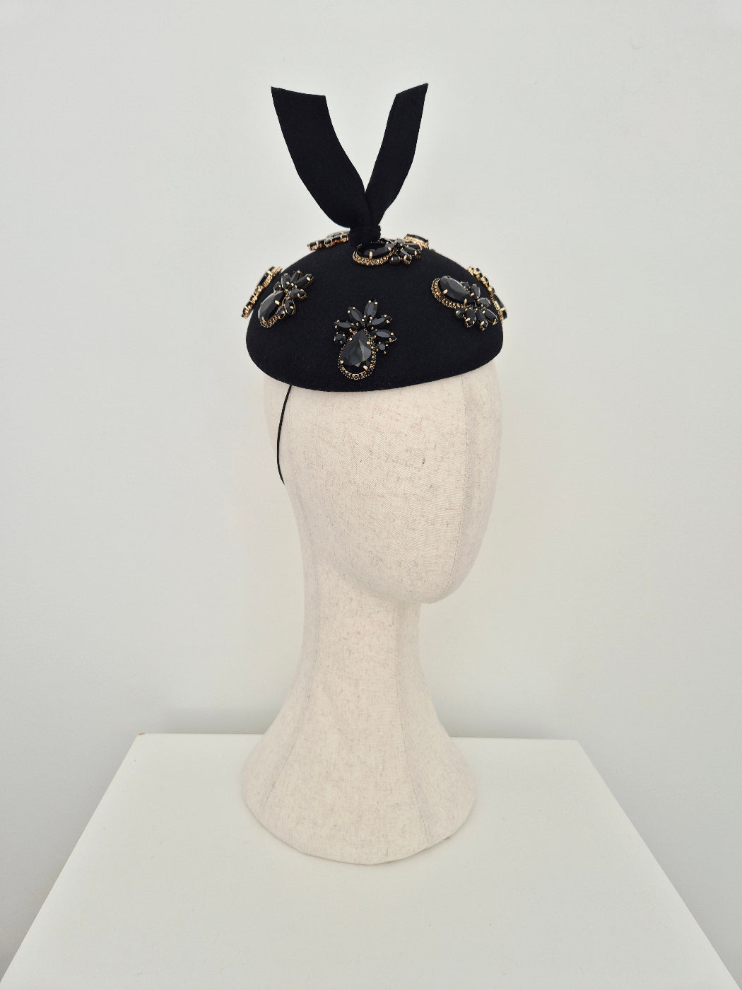 A black felt hat with rhinestone detail and a black ribbon bow, displayed on a cream-colored felt mannequin head against a minimalist white background. side view