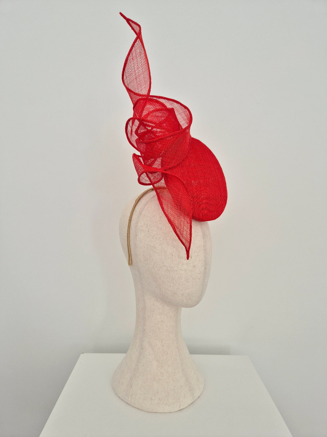 A bright red fascinator hat displayed on a textured off-white mannequin head. The hat features a sculptural, flowing red mesh design with a large rounded base and a dramatic upward curl.side view