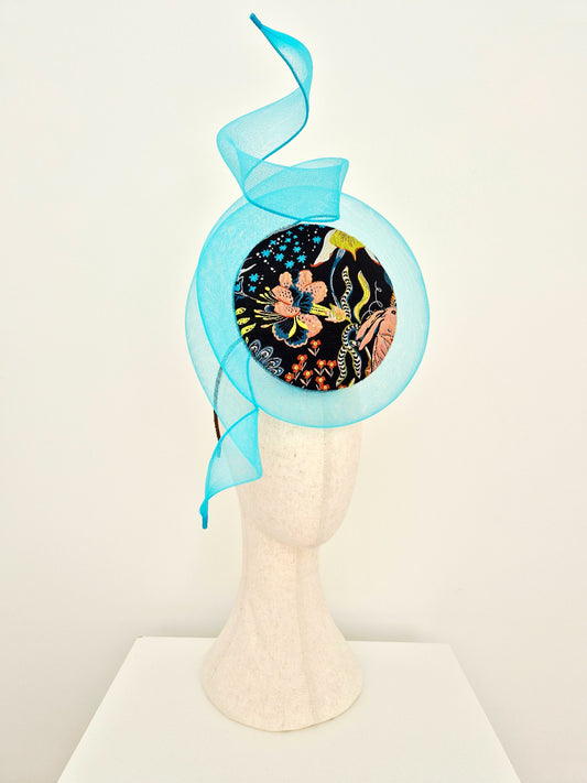 A blue crinoline swirl and tails fascinator  with a circular black floral patterned center, displayed on a textured off-white mannequin head against a plain white background.side view