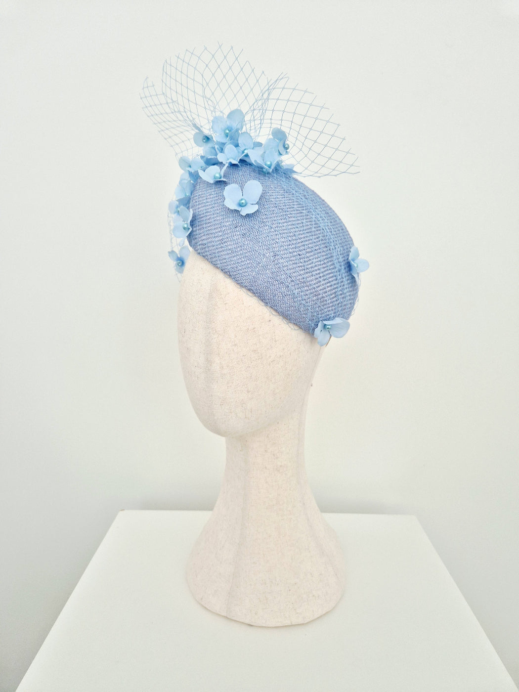 A light blue woven fascinator with light blue fabric flowers and a netted veil, displayed on a textured off-white mannequin head against a plain white background.side view