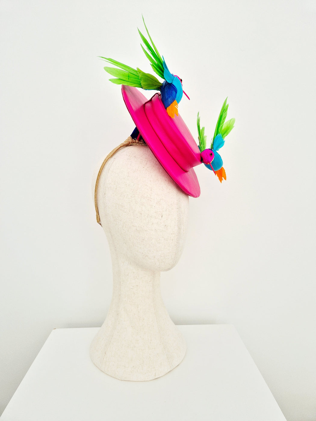 A bright pink fascinator hat displayed on a textured off-white mannequin head. The hat features a wide pink brim and is decorated with two colorful bird embellishments in blue, green, and orange, set against a plain white background. side view