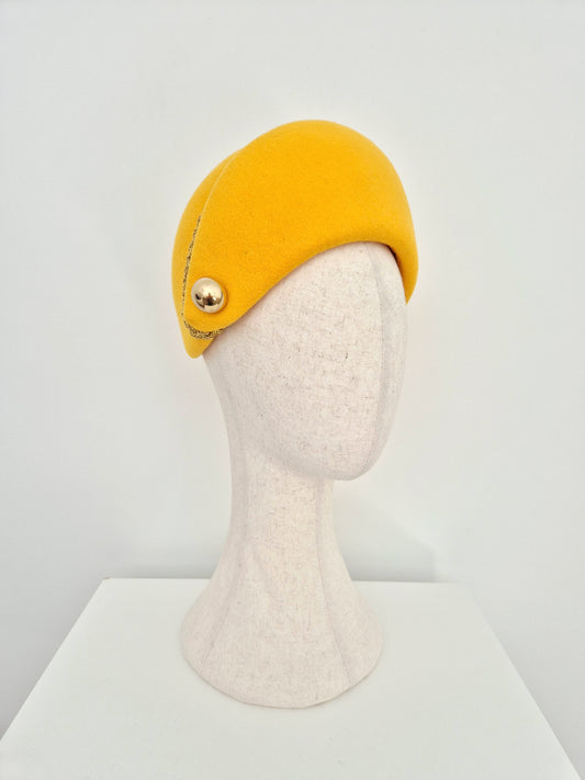 A yellow felt hat on a mannequin head, featuring a decorative gold button on the side, set against a plain white background.