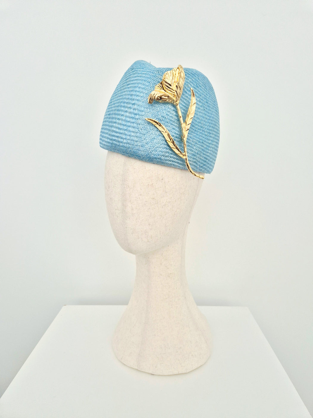 A sky blue straw touque headpiece displayed on a cream-colored mannequin head. The hat features a prominent gold metal flower and leaf embellishment on the side, set against a clean white background.