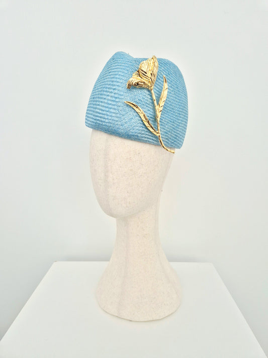 A sky blue straw touque headpiece displayed on a cream-colored mannequin head. The hat features a prominent gold metal flower and leaf embellishment on the side, set against a clean white background.