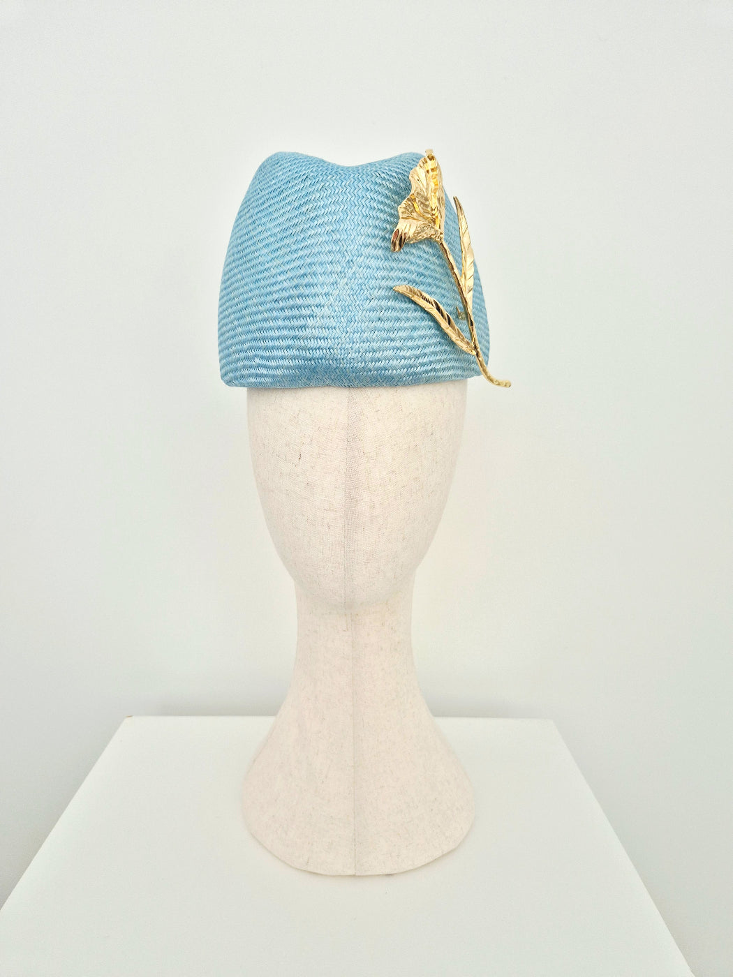 A sky blue straw fascinator headpiece with a gold flower accent, displayed on a cream-colored mannequin head against a white background.