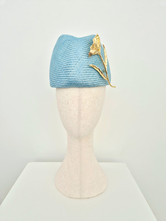 A sky blue straw fascinator headpiece with a gold flower accent, displayed on a cream-colored mannequin head against a white background.