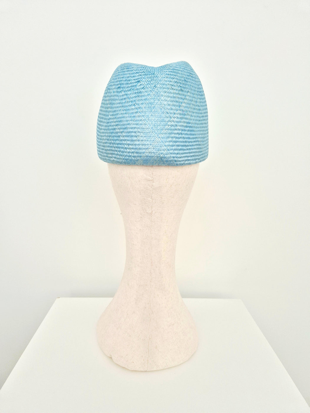 A sky blue straw touque headpiece displayed on a cream mannequin head stand against a clean white background.back view