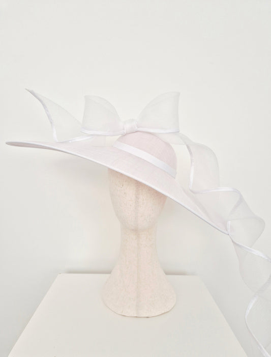 A white fascinator hat with a wide brim, sheer white crinoline bow, and long trailing ribbon, displayed on a textured cream mannequin head against a white background.