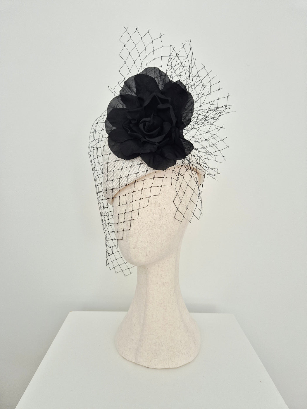 A white straw fascinator with a large fabric rose and matching black birdnet veil, displayed on a cream-colored felt mannequin head against a plain white background.side view