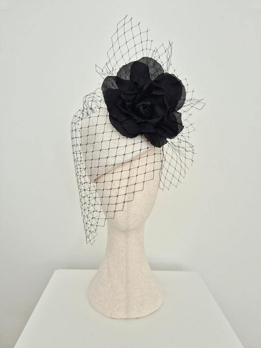 A white straw fascinator with a large black rosette flower and a long black net veil, displayed on a cream-colored mannequin head against a white background.