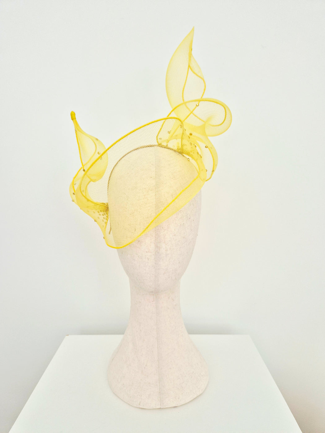 A bright yellow crinoline fly away design fascinator hat with scattered rhinestones displayed on a textured off-white mannequin head against a clean white background