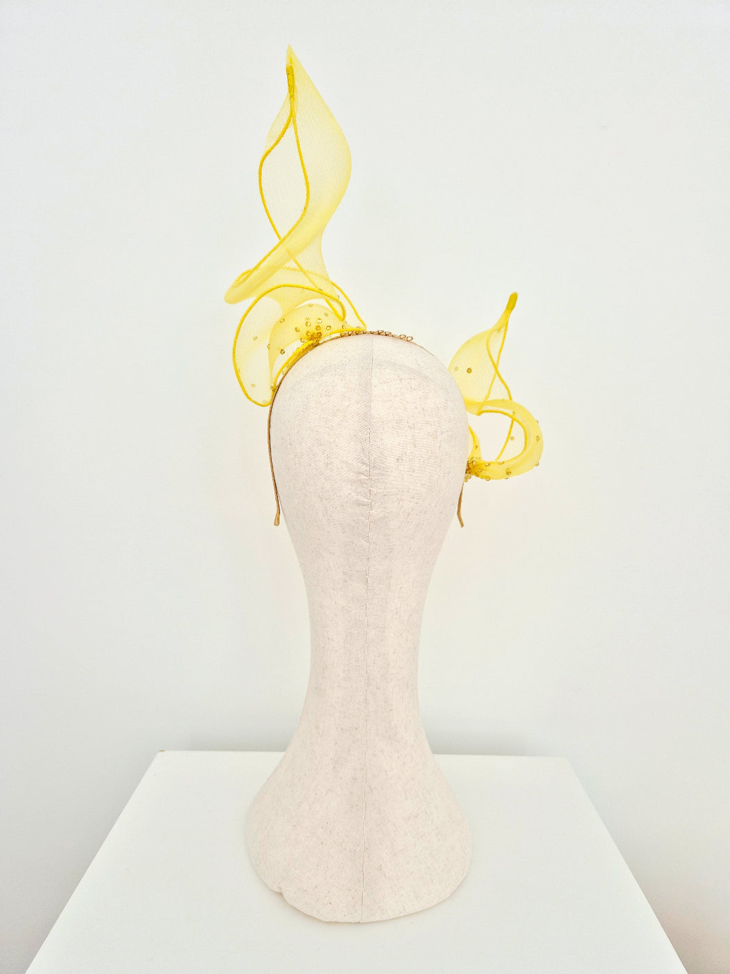 A mannequin head wearing a dramatic yellow fascinator headpiece with flowing sheer leaf-like loops, photographed against a clean white studio background.back view